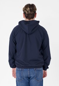 Giacca a vento blu navy con cappuccio, polsini e vita elasticizzati, mostrata da dietro, abbinata a jeans azzurri chiari.