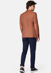 Brązowa marynarka w tweed z brązowymi skórzanymi łokciami, połączona z granatowymi spodniami i czarnymi sneakersami. Dopasowany krój, teksturowany materiał i zdefiniowana sylwetka.