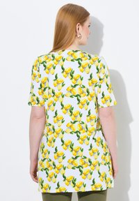 Tunic bianca con motivo di limoni e foglie, maniche corte e vestibilità ampia. La vista posteriore mostra colori vivaci e dettagli di design chiari.