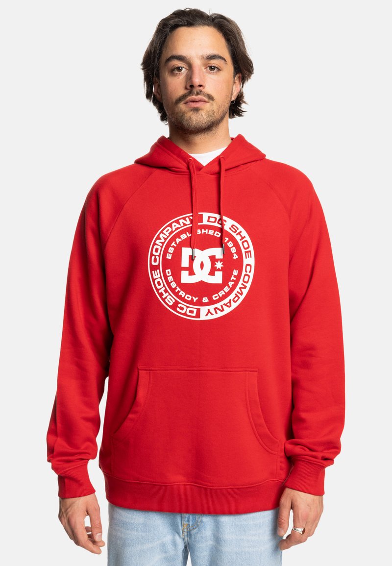 Red hoodie with a large white circular logo featuring "DC Shoe Company" text. Soft cotton material, kangaroo pocket, ribbed cuffs, and hem.