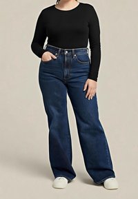 Femme portant un top noir à manches longues, un jean bleu foncé taille haute à jambes larges et des chaussures blanches, une main dans la poche et l’autre reposant sur la cuisse.
