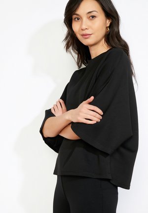 Žena s dlhými tmavými vlasmi, oblečená v čiernom oversized topánke a čiernych nohaviciach, stojaca so založenými rukami na bielom pozadí.