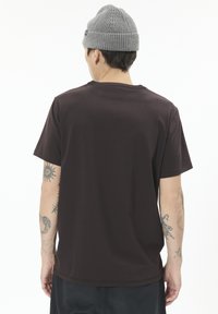 Hnědá košile s krátkým rukávem a kulatým výstřihem, hladká textilie a uvolněný střih, nošena s šedou pletenou čepicí, tetování viditelné na předloktích.