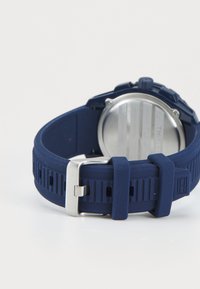 Montre bleue en silicone avec un bracelet texturé, boucle en métal et boîtier circulaire en argent. Présente un dos simple avec des lignes horizontales.