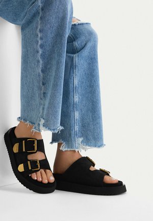 Piedi che indossano sandali piattaforma neri con fibbie dorate, abbinati a jeans blu chiaro con orlo sfrangiato su uno sfondo bianco semplice.