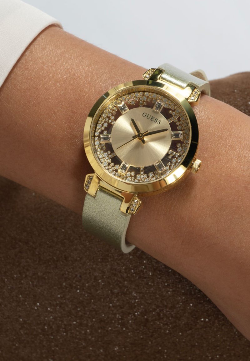 Orologio dorato con quadrante rotondo, decorazioni in cristallo e cinturino in pelle verde chiaro. Presenta un design elegante e indici delle ore minimalisti.