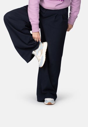 Persoon die een marineblauwe broek met wijde pijpen en lichtblauwe sneakers draagt, tilde een been op en houdt het broekspijp vast om de schoen te laten zien.