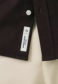 Gros plan sur l'ourlet d'une chemise marron foncé avec une étiquette blanche indiquant "Durable Goods LOT.082" et un bouton blanc au-dessus.