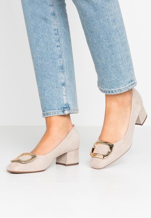 Fødder iført lyse beige ruskindsblokhæle med store guldspænder, parret med lyseblå cropped denim jeans.