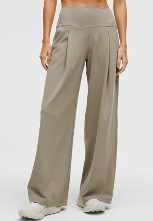 Woman wearing high-waisted, wide-leg taupe pants paired with white sneakers, standing against a plain light background.