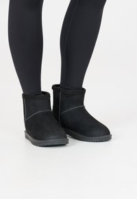 Botines de ante negros con una superficie texturizada, forro suave y una suela de goma resistente. Presentan costuras decorativas a lo largo de los costados.