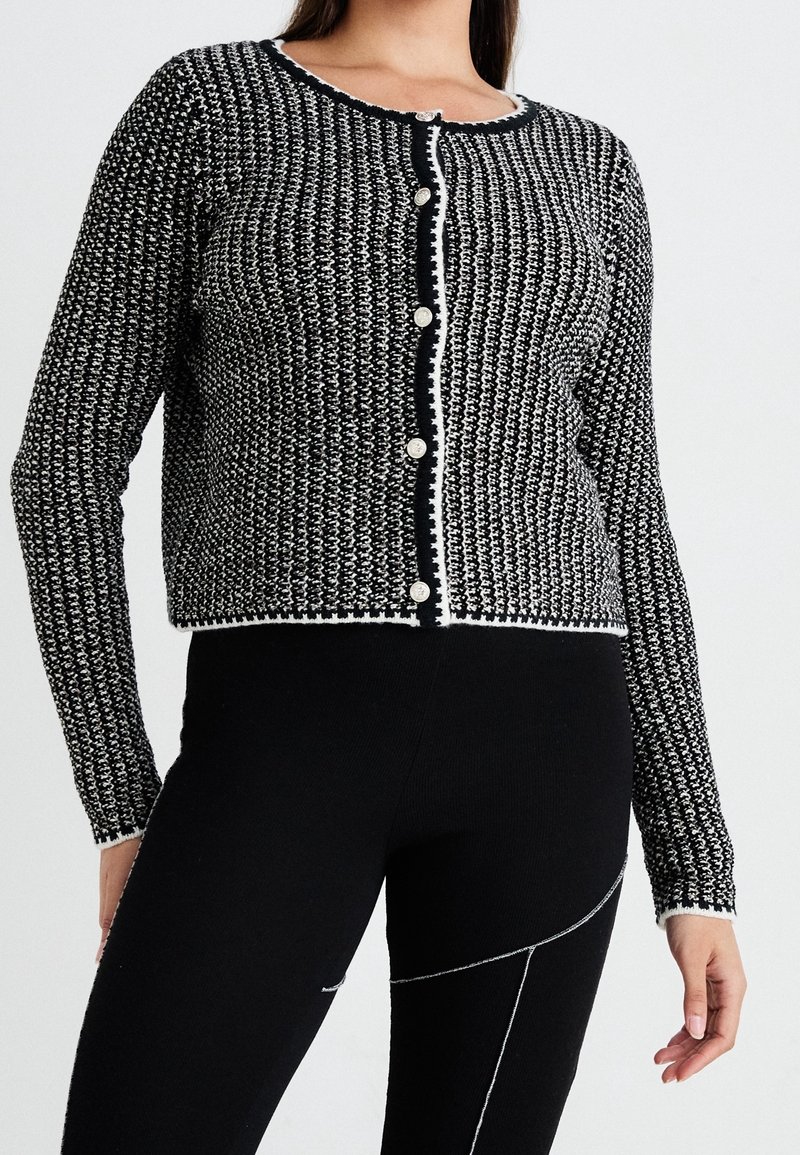 Cardigan à motifs noir et blanc avec un col rond, des boutons et une bordure contrastante. Manches longues et coupe courte.