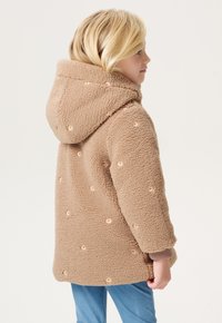 Beige Fleecejacke mit Kapuze, die über kleine floral embroiderte Details, eine strukturierte Oberfläche und gerippte Bündchen verfügt. Sie wird über blauen Jeans getragen.