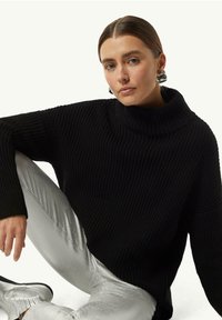 Schwarzer Strickpullover mit hohem Kragen und Rippstruktur, kombiniert mit glänzenden silbernen Hosen. Das Modell posiert sitzend und hebt das Design des Outfits hervor.