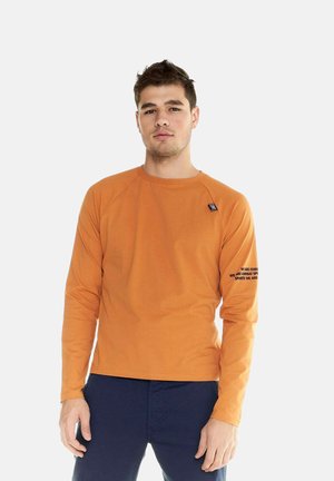 Mann trägt ein oranges Langarmshirt mit schwarzem Text auf dem linken Ärmel und dunkle Hose, steht vor weißem Hintergrund.