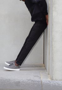 Czarne wąskie spodnie w połączeniu z szarymi zamszowymi sneakersami z białymi podeszwami. Strój na tle teksturowanej, szarej ściany.