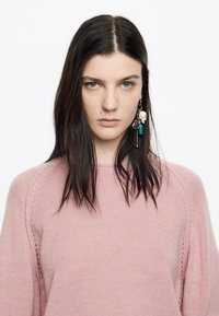 Maglione lavorato a maglia rosa chiaro con scollatura rotonda e dettagli a coste ai bordi. Maniche lunghe, texture morbida, indossato con orecchini decorativi.
