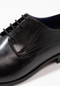 Chaussure habillée en cuir noir avec une surface lisse, un bout rond et un système de laçage, ornée de coutures discrètes et d'un logo embossé.