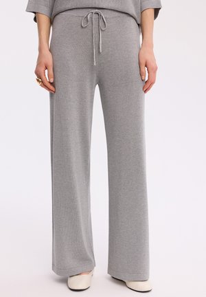 Kobieta nosząca jasnoszare szerokie spodnie dzianinowe z wiązaniem w talii i białe płaskie buty, stojąca na jednolitym tle.