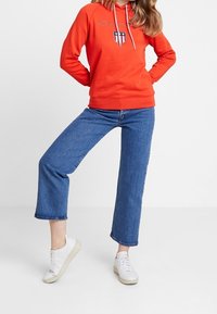 Hoodie rouge avec logo sur la poitrine, associé à un jean en denim bleu à jambes larges et des baskets blanches. Le hoodie présente un cordon de serrage et des poignets côtelés.