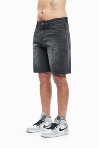 Mężczyzna w czarnych szortach dżinsowych z haftowanymi wzorami oraz białych wysokich butach Nike z czarnymi akcentami, stojący na białym tle.