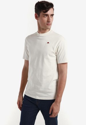 Joven con una camiseta blanca lisa de manga corta con un pequeño logo rojo en el pecho, lleva vaqueros azul oscuro, de pie frente a un fondo blanco.