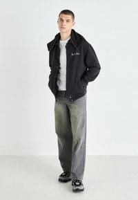 Svart, knappet jakke med dobbel-lags hette, kombinert med grå jeans. Skoene har et strukturert svart og hvitt design. En enkel hvit skjorte under.