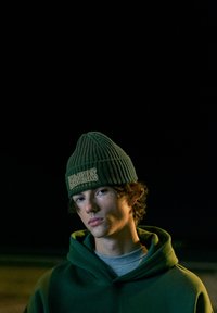 Beanie ribbed verde cu logo-ul "HAWKINS", asociat cu un hanorac supradimensionat asortat. Textură moale, design cald și fundal întunecat.