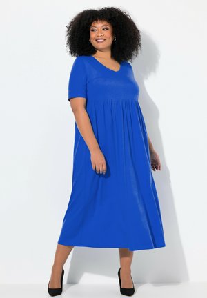 Robe maxi bleu à col en V avec des manches courtes, corsage ajusté et jupe évasée. Tissu lisse, portée avec des talons noirs.