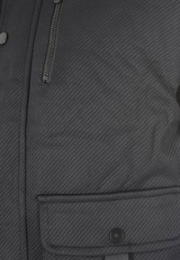Giacca nera con texture, dotata di chiusura con zip, tasca frontale con pattina e dettaglio a bottone rotondo. Tessuto liscio con righe diagonali.