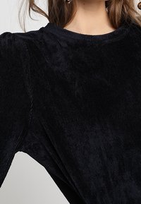 Svart kordflodertopp med en texturerad yta, med rund hals och lätt puffärm, som framhäver tygets djup och mjukhet.