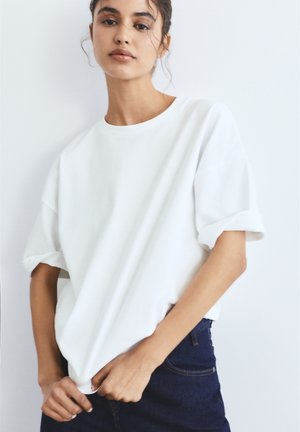 Junge Frau mit dunklen Haaren in einem weißen, übergroßen T-Shirt und dunklen Jeans, die den Saum an einer schlichten weißen Wand anpasst.