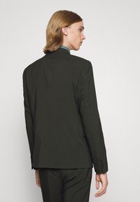 Dunkelgrüner, maßgeschneiderter Anzugjacke mit einer einzelnen Rückenschlitz, eingekerbten Revers und Knopfakzenten an den Ärmeln. Glatte Stofftextur.