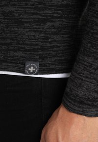 Pull gris texturé avec un motif tissé, comportant une étiquette avec un logo en croix sur l'ourlet. Superposé à un t-shirt blanc. Jeans noirs visibles.