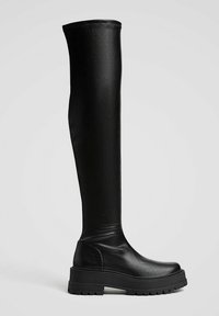 Botte noire unique au-dessus du genou avec semelle à crampons épaisse et bout arrondi, en cuir lisse ou matériau similaire sur un fond clair uni.