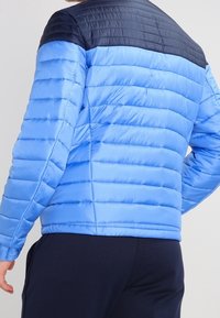 Vadderad jacka med en klarblå kropp och marinblå axelparti, med ett quiltat mönster och slät textur.