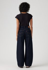 Traperice širokih nogavica visokog struka, tamnoindigo boje, s kožnim detaljem i dvostrukim stražnjim džepovima, uparene s crnom majicom s kratkim rukavima.