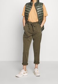 Person, der olivgrüne hochtaillierte Paperbag-Hosen, beigen Sneaker, ein orangefarbenes Kurzarmhemd und eine dunkelgrüne gesteppte Weste trägt, steht mit den Händen in den Taschen.