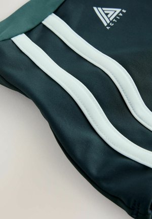 Tumedrohelised sportlikud lühikesed püksid, millel on kontrastsed helesinised triibud ja väike logo. Kangas tundub sile ja kergelt läikiv.