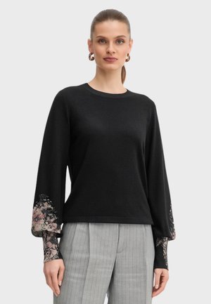 Maglione nero lavorato a maglia con maniche lunghe e dettagli in pizzo floreale ai polsini. Abbinato a pantaloni grigi a righe.