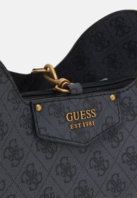 Zwarte handtas met textuur, voorzien van een gouden "GUESS EST 1981"-logo en decoratieve hardware. Het oppervlak heeft een subtiel patroon.