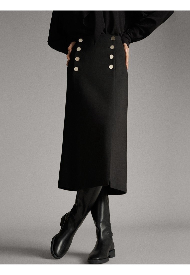 Femme portant une jupe midi noire avec des boutons argentés et des bottes noires montant jusqu’aux genoux, debout les mains sur les hanches devant un fond uni.