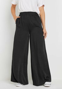 Pantalon large noir en tissu léger, avec une taille élastique et des poches latérales. La coupe offre une silhouette décontractée.
