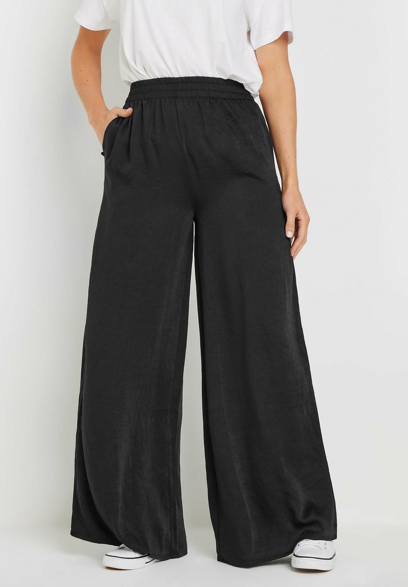 Pantalon large noir en tissu léger, avec une taille élastique et des poches latérales. La coupe offre une silhouette décontractée.