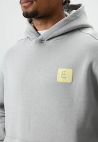 Grijze hoodie met een zachte textuur, voorzien van een vierkant geel patch met een gestileerde letter "E" en een steraccent aan de voorkant.