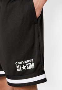 Main d'une personne dans la poche d'un short Converse All Star noir avec des bandes blanches près de l'ourlet sur un fond uni.
