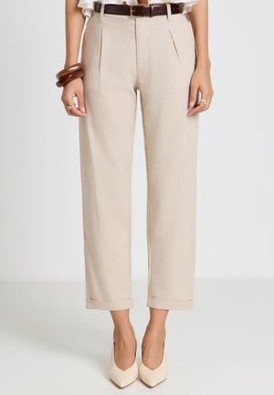 Beige hoch taillierte, maßgeschneiderte Hose mit umgeschlagenen Säumen, kombiniert mit einem braunen Gürtel, weißen spitzen Absätzen und Armband-Accessoires an einem Model.