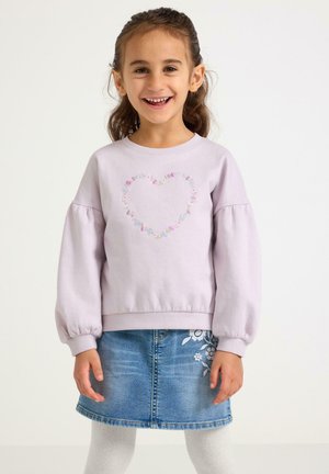 Ragazza sorridente con capelli castani, indossa una felpa viola chiaro con un cuore floreale e una gonna di jeans blu con ricami floreali.