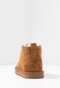 Bottine UGG en daim marron avec languette de tirage et doublure en peau de mouton visible, vue de dos sur une surface blanche avec un coin de mur en arrière-plan.