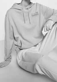 Sudadera gris claro de tejido suave con cordones, bolsillo frontal y detalle de logo impreso. Combinada con pantalones gris claro a juego.